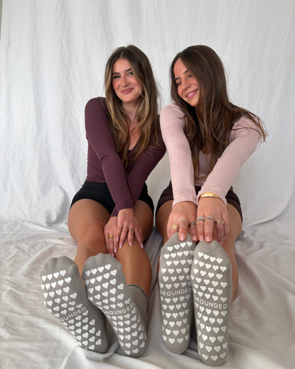 Two women wearing gray ruffle pilates socks with heart patterns and the brand name 'GROUNDED'.