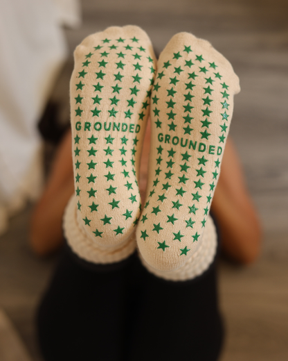 Beige grippy socks with green star pattern and 'Grounded' text worn by a person.